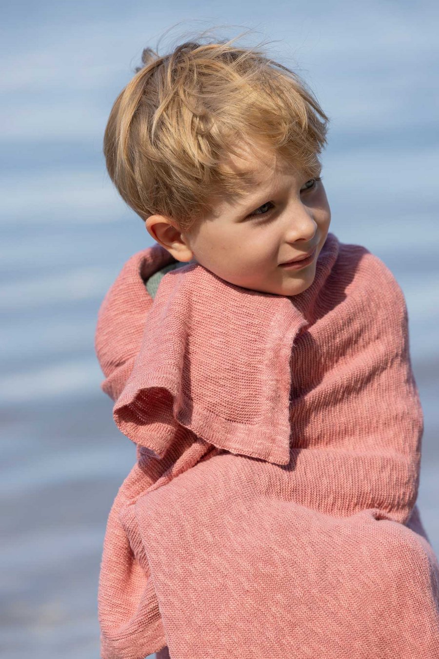 Ein blonder Junge hat sich in einer disana Leinen-Decke, Softpink eingerollt & schaut nach rechts. Hintergrund ein blauer See