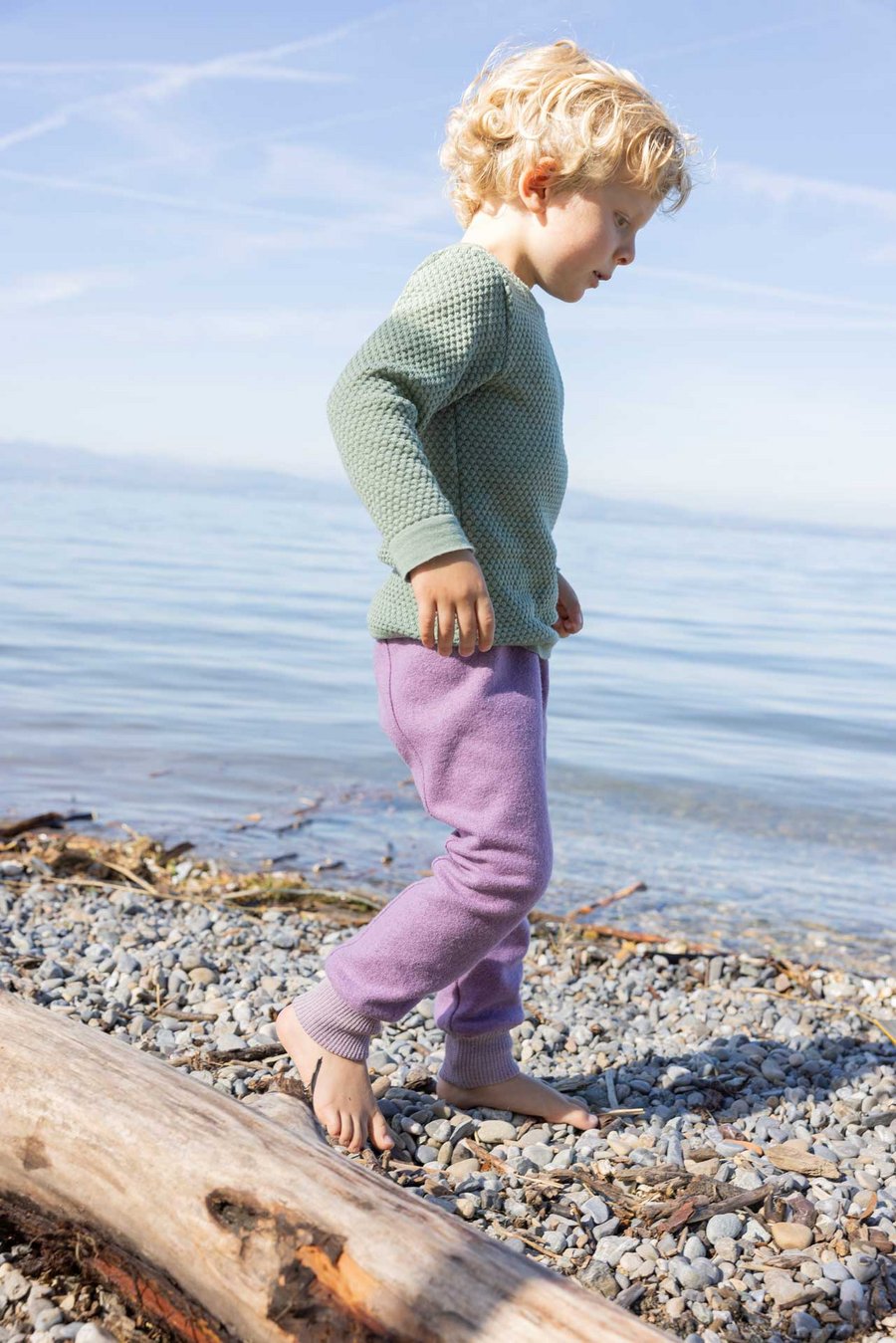 Blondes Kind in disana Wabenstrick-Pulli, Jade & Pumphose, Flieder läuft nach rechts am Seeufer - Wolle aus Bio-Merinowolle