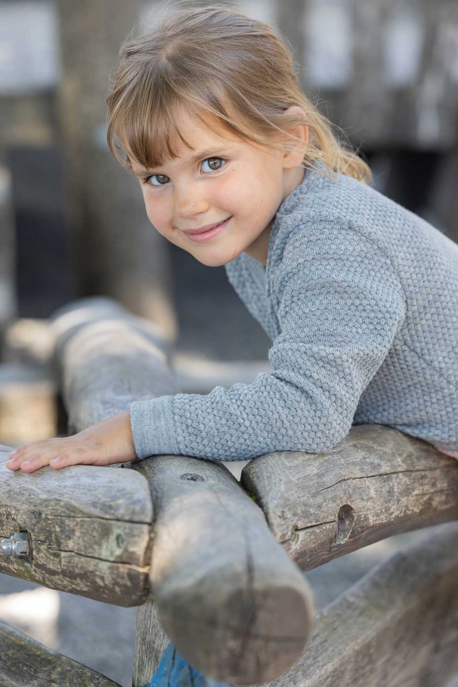 Kind in grauem disana Wabenstrick-Pullover spielt draußen & lehnt sich nach vorne auf Holzstämme, Strick aus Bio-Merinowolle