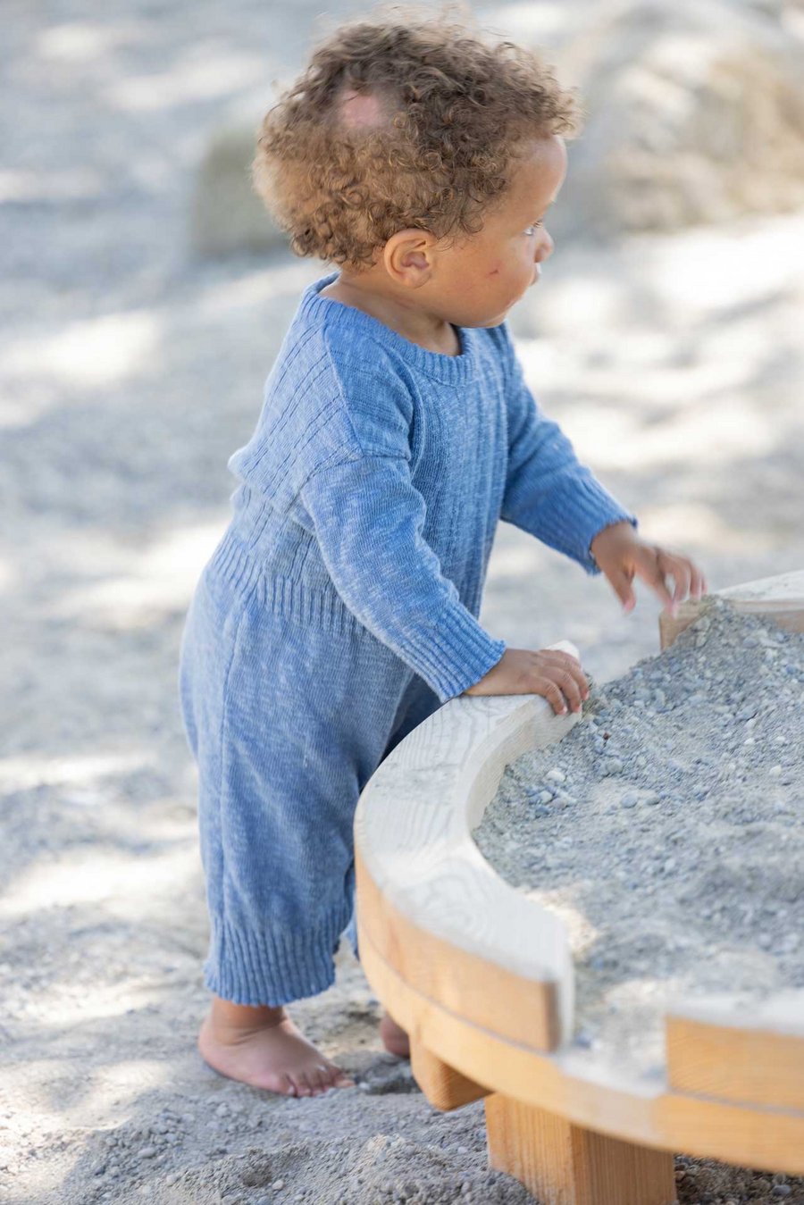 Dunkelhäutiges Kleinkind trägt disana Leinen-Strampler, Taubenblau und spielt barfuss im Sandkasten. Es schaut nach links