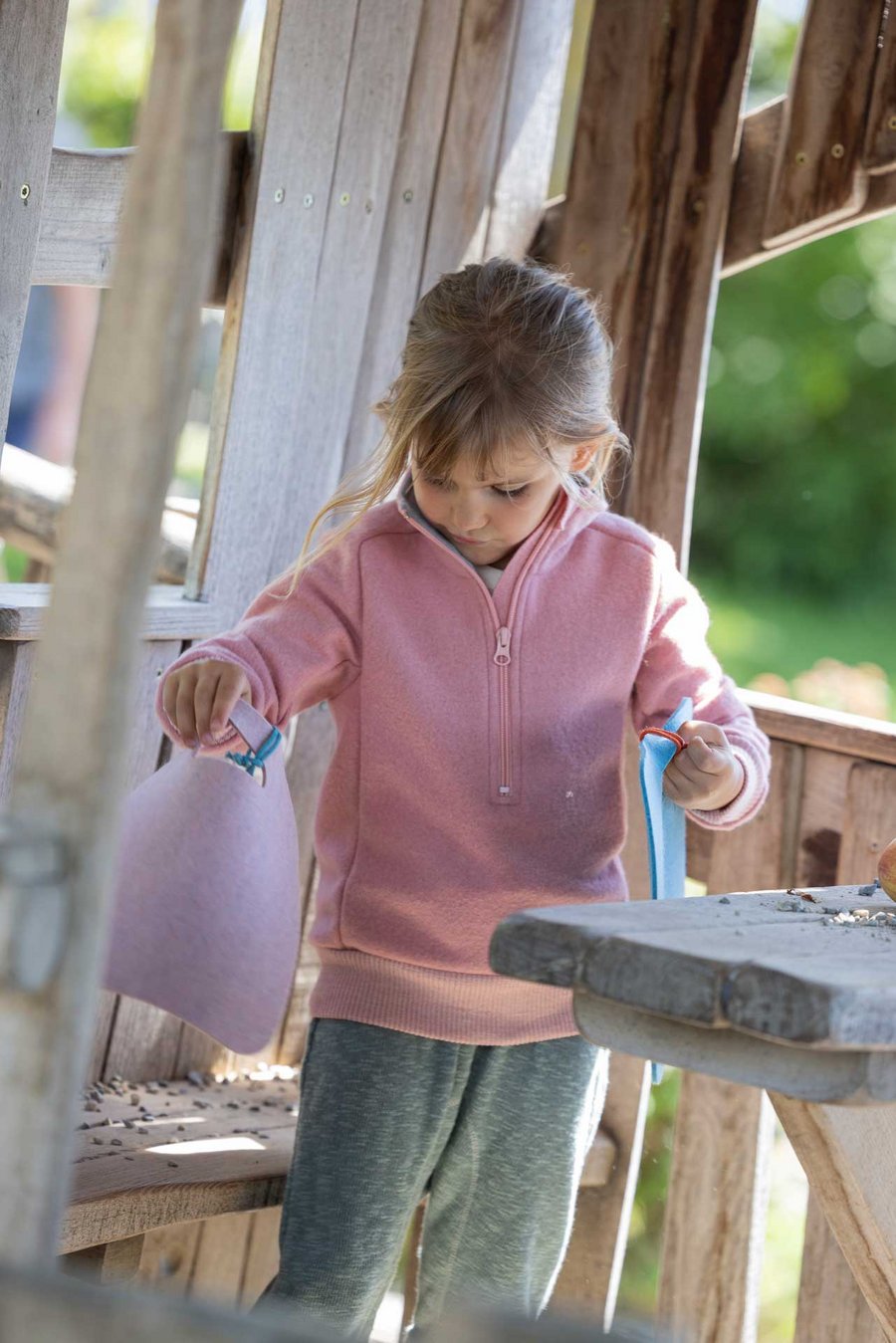Mädchen im disana Troyer Softpink mit Leinen-Strickhose in Jade hält zwei Filz-Sitz-Auflagen Softpink & Lagoon in jeder Hand.