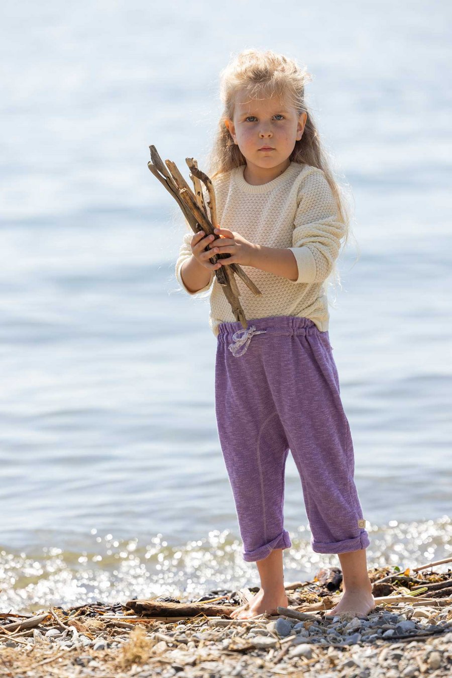 Mädchen mit langen blonden Haaren, im disana Waben-Pulli Natur & Leinen-Strickhose Flieder steht am Seeufer & hält Holzstäben