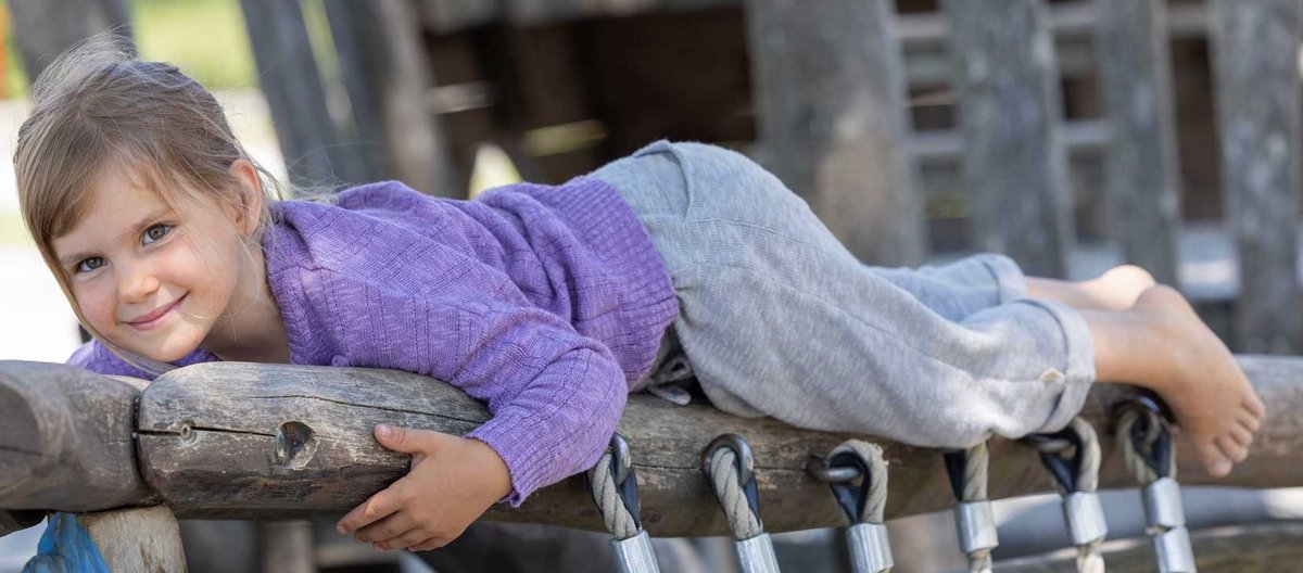 Mädchen in fliederfarbenem disana Leinenpulli & grauer Leinen-Strickhose auf einem verwitterten Klettergerüst liegend