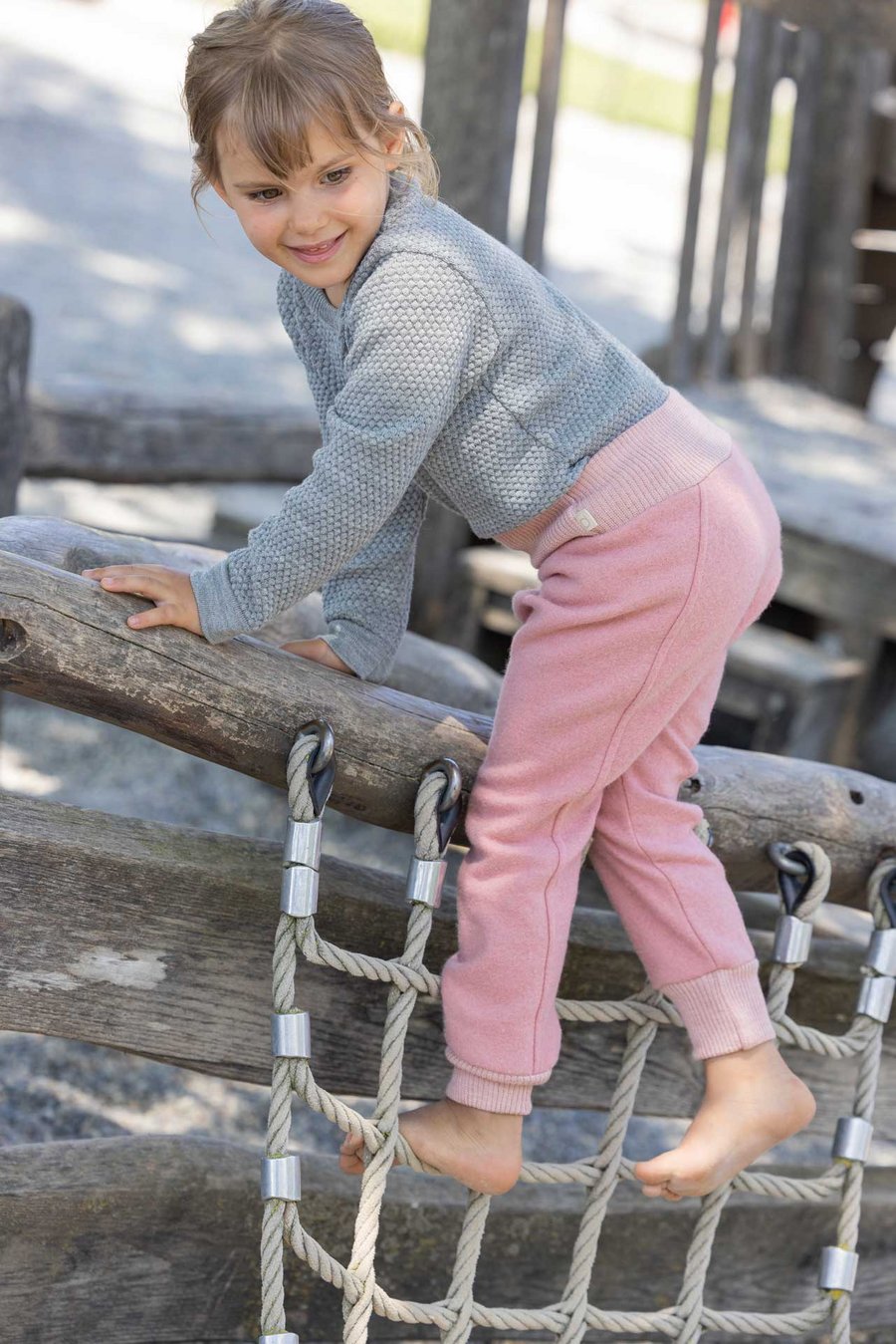 Kind goldbraune Haare trägt disana Wabenstrick-Pulli Grau & Pumphose Softpink auf Kletternetz und schaut nach unten links