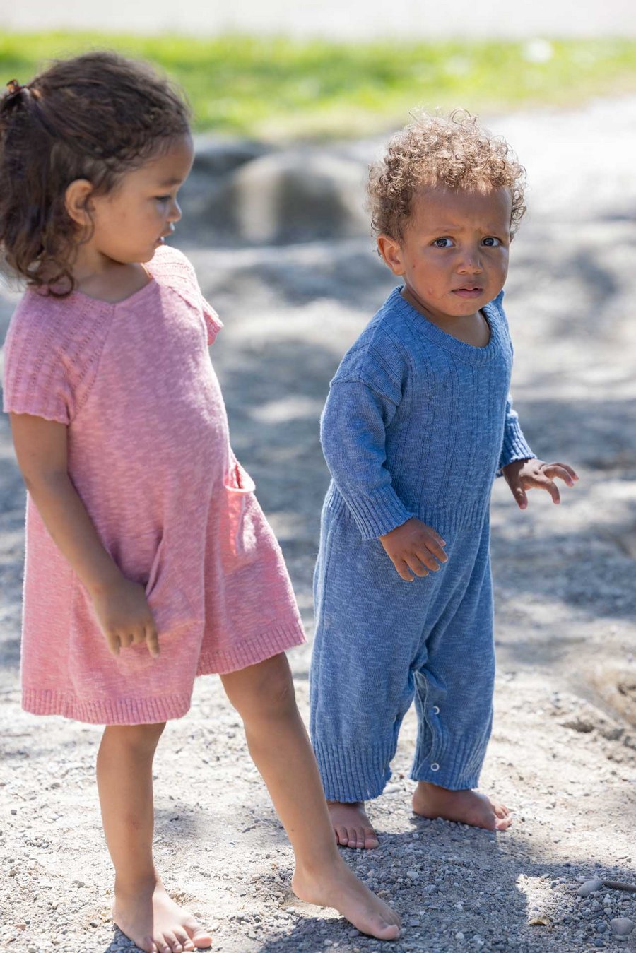 Zwei dunkelhäutige Kinder barfuss im Sand. Das jüngste im disana Strampler, Taubenblau, das Mädchen im einenkleid, Softpink