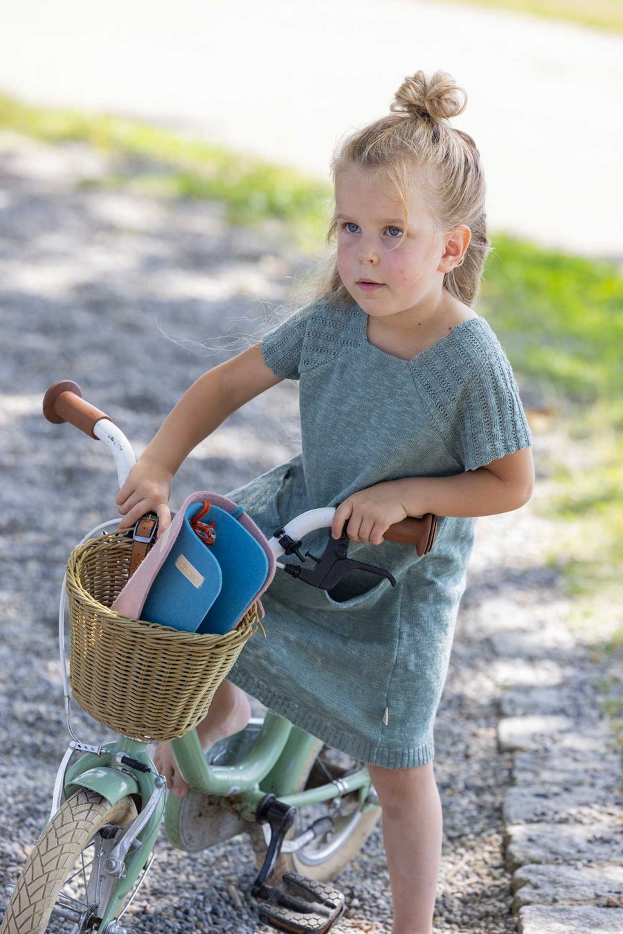 Ein blondes Mädchen mit Halfbun & einem disana Leinen-Kleid, Jade, sitzt auf einem hellgrünen Rad mit einem Korb am lenkrad