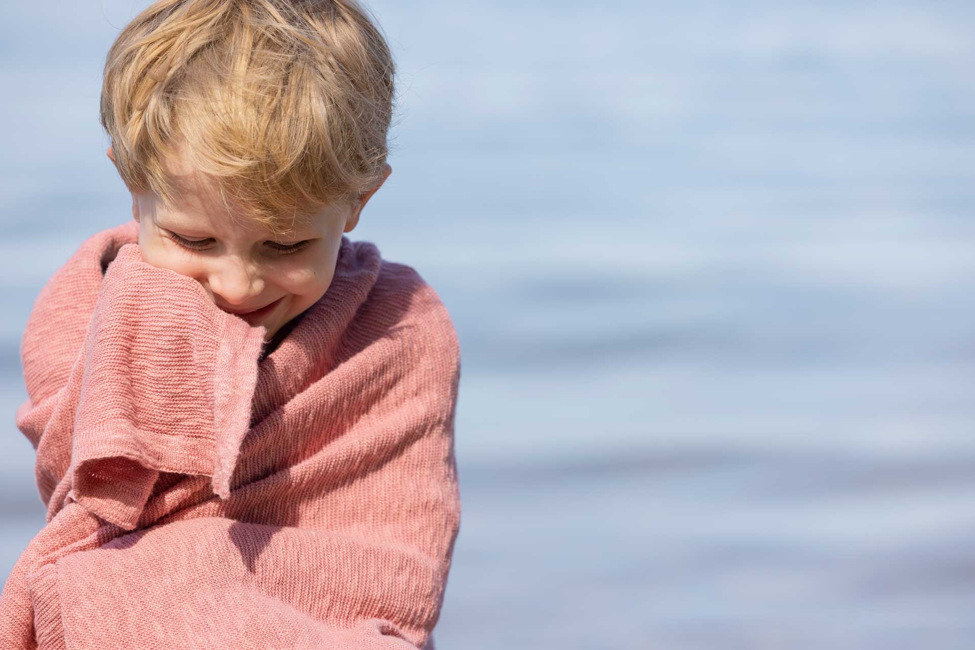 Blonder Junge eingekuschelt in einer softpinkfarbenen disana Leinen-Decke aus Bio-Baumwolle-Leinen-Mix vor See-Hintergrund.