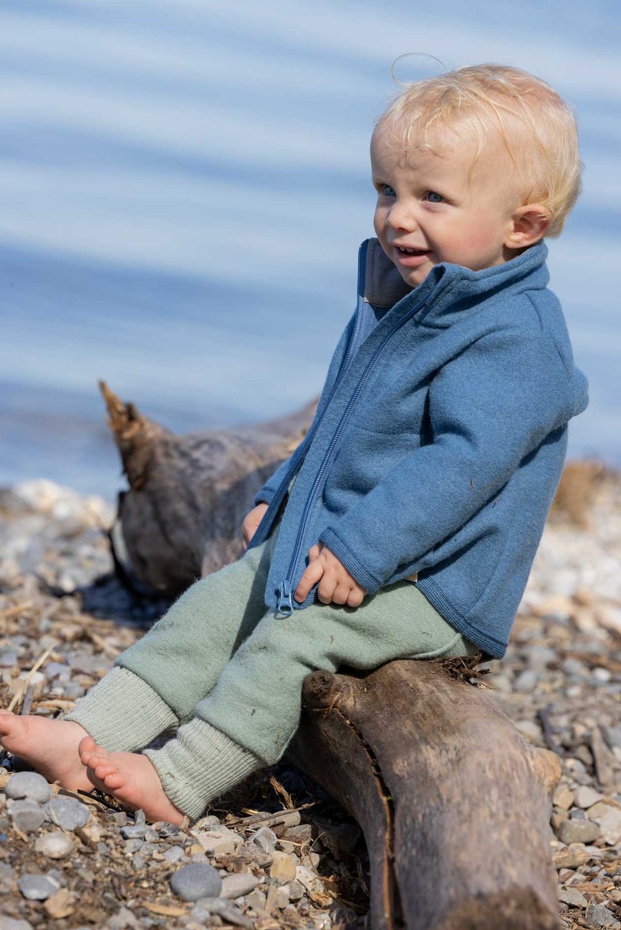 Kleinkind in disana Zipper-Jacke Taubenblau & Pumphose in Jade an einem Seeufer auf einem Holzstamm sitzend - Sommer-Wollwalk