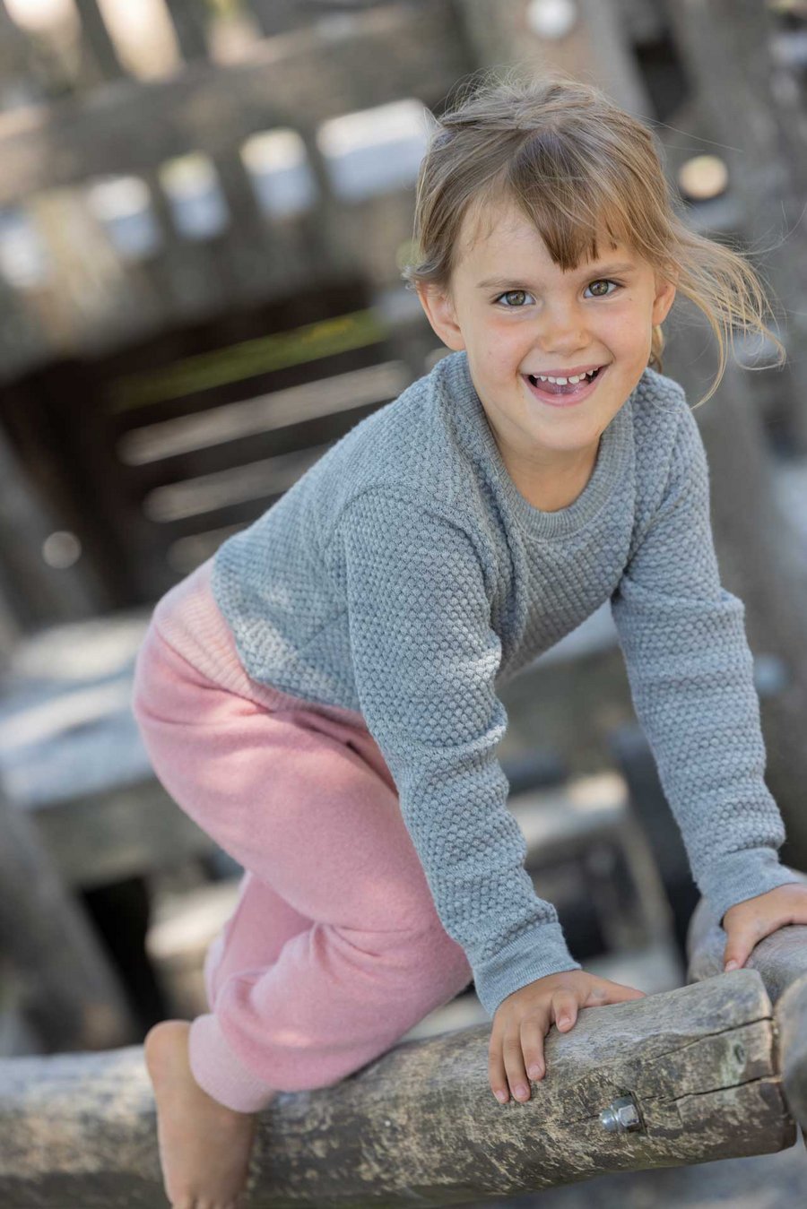Kind goldbraune Haare trägt disana Wabenstrick-Pulli Grau & Pumphose Softpink auf Holzgerüst kletternd lacht in die Kamera