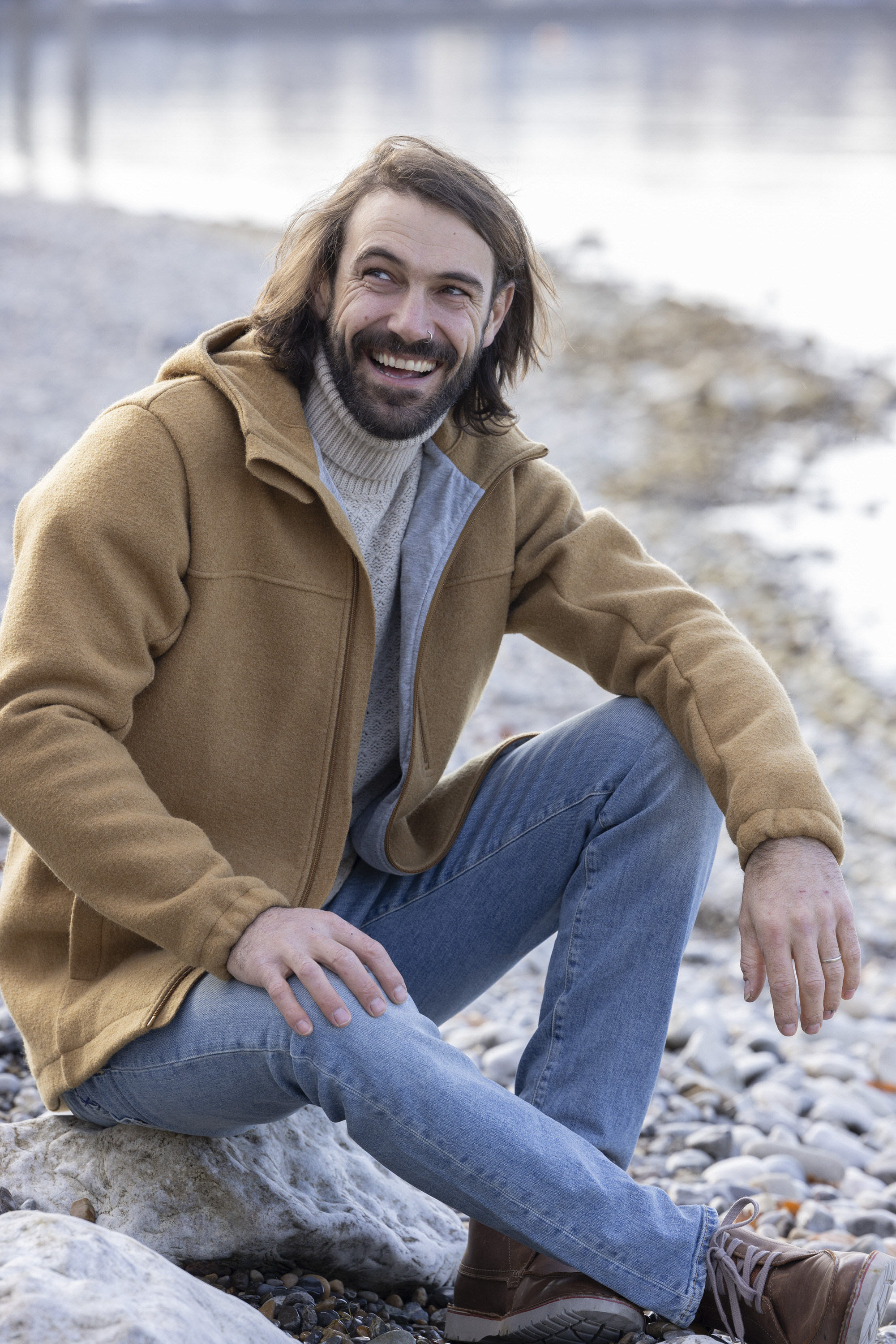 Lächelnder Mann mit dunkelbraunen Haaren in disana Herren-Jacke Toffee und Jeanshose sitzend an einem Seeufer