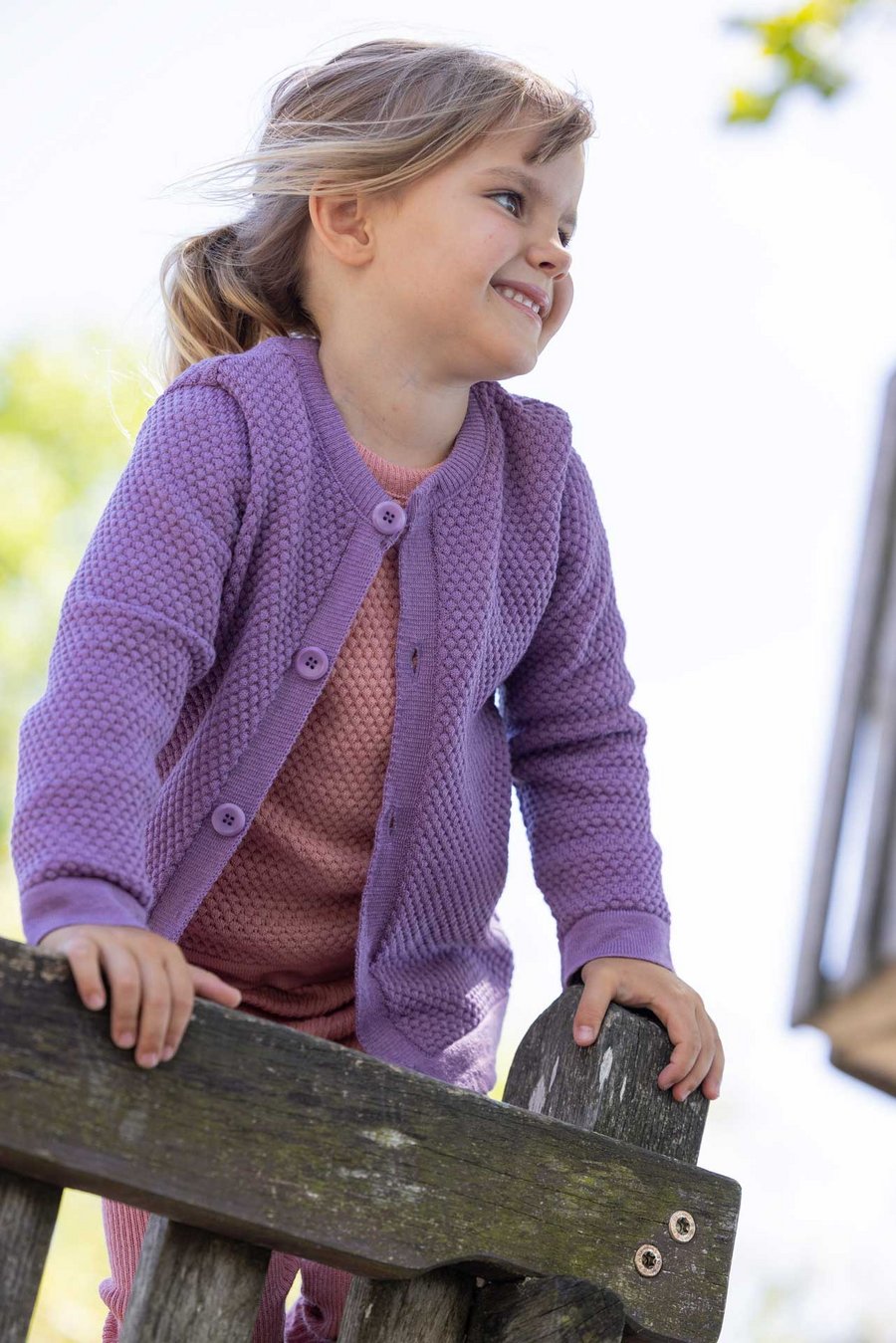 Kind trägt fliederfarbene disana Waben-Strickjacke offen aus Bio-Merinowolle, lachend,  spielt auf einem Holz-Klettergerüst