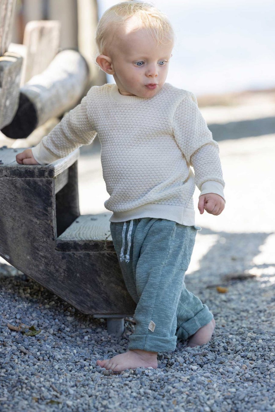 Kleinkind im disana Waben-Pulli, Natur & Leinen-Strickhose, Jade steht barfuß auf Kiesweg neben einer verwitterten Holztreppe