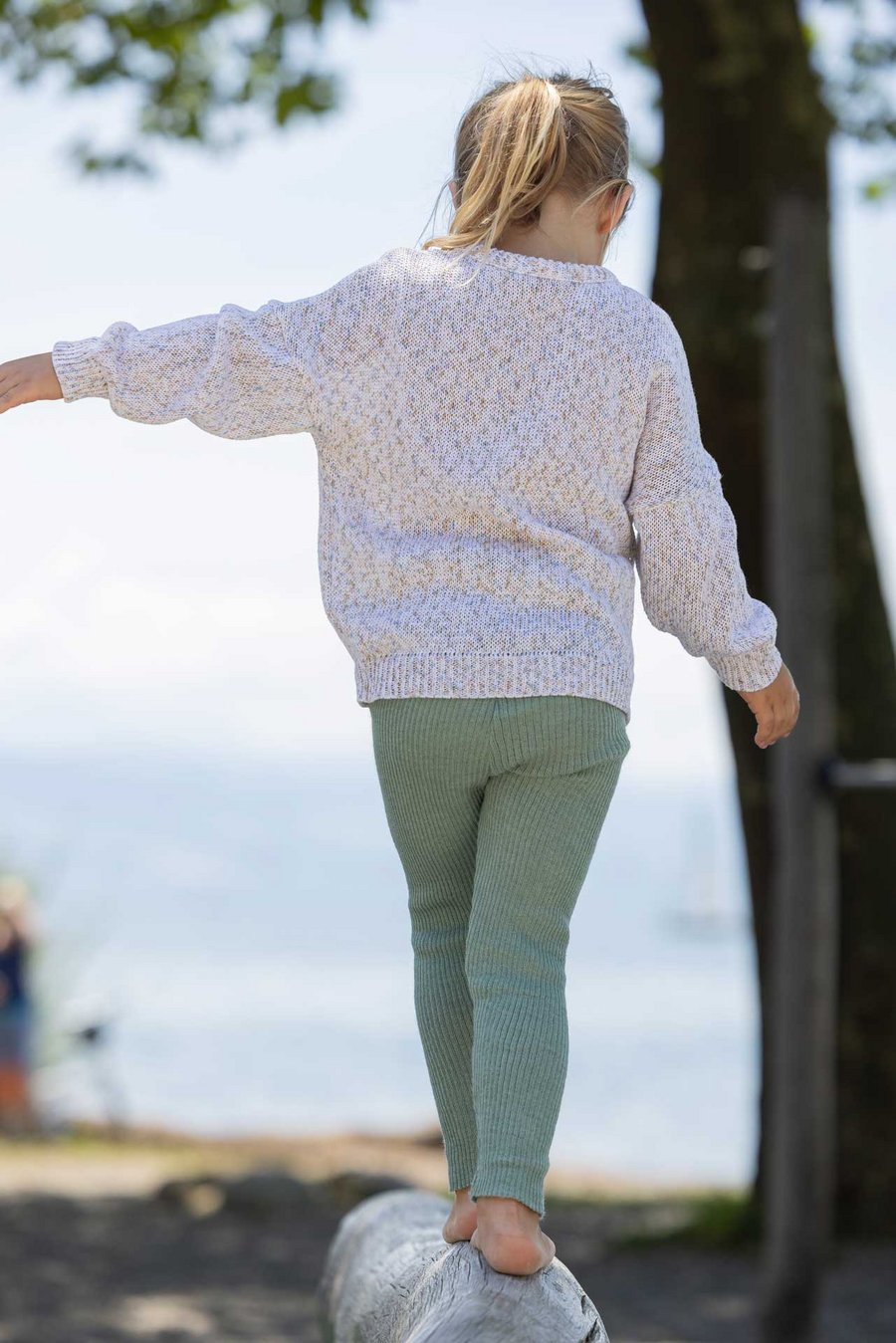 Blondes Mädchen mit Pferdeschwanz auf Holzstamm balancierend. Im disana Baumwoll-Pulli Spray, Natur & Leggings-Light Jade