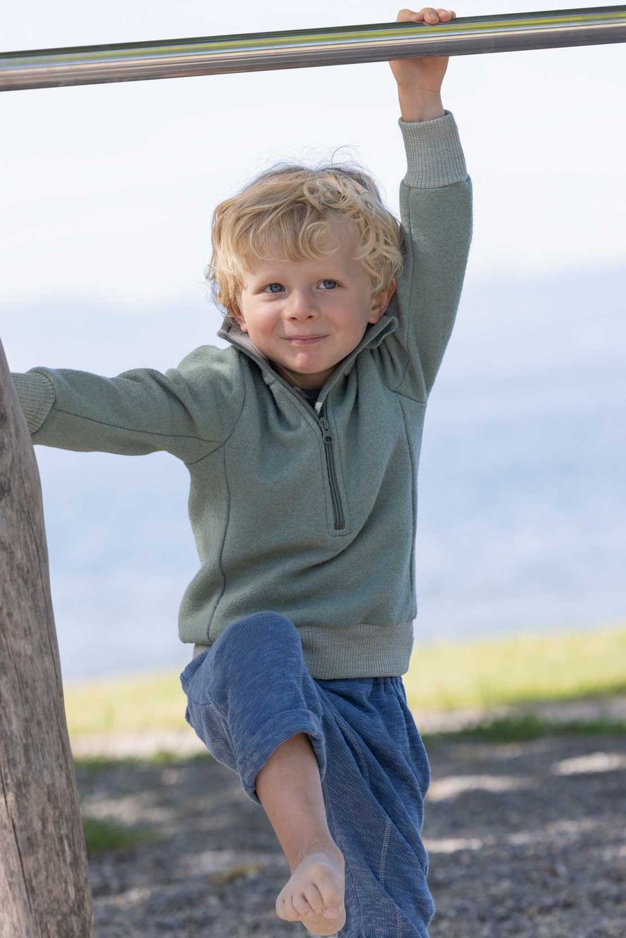 Blonder Junge im disana Troyer in Jade und Leinen-Strickhose in Taubenblau, spielt an einer Turnstange am Seeufer