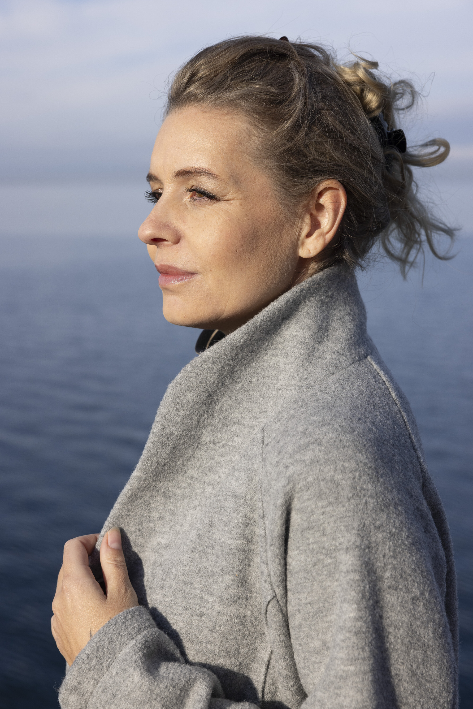 Seitenprofil einer blonden Frau mit hochgesteckten Haaren im grauen disana Damen-Mantel am Seeufer an einem sonnigen Tag