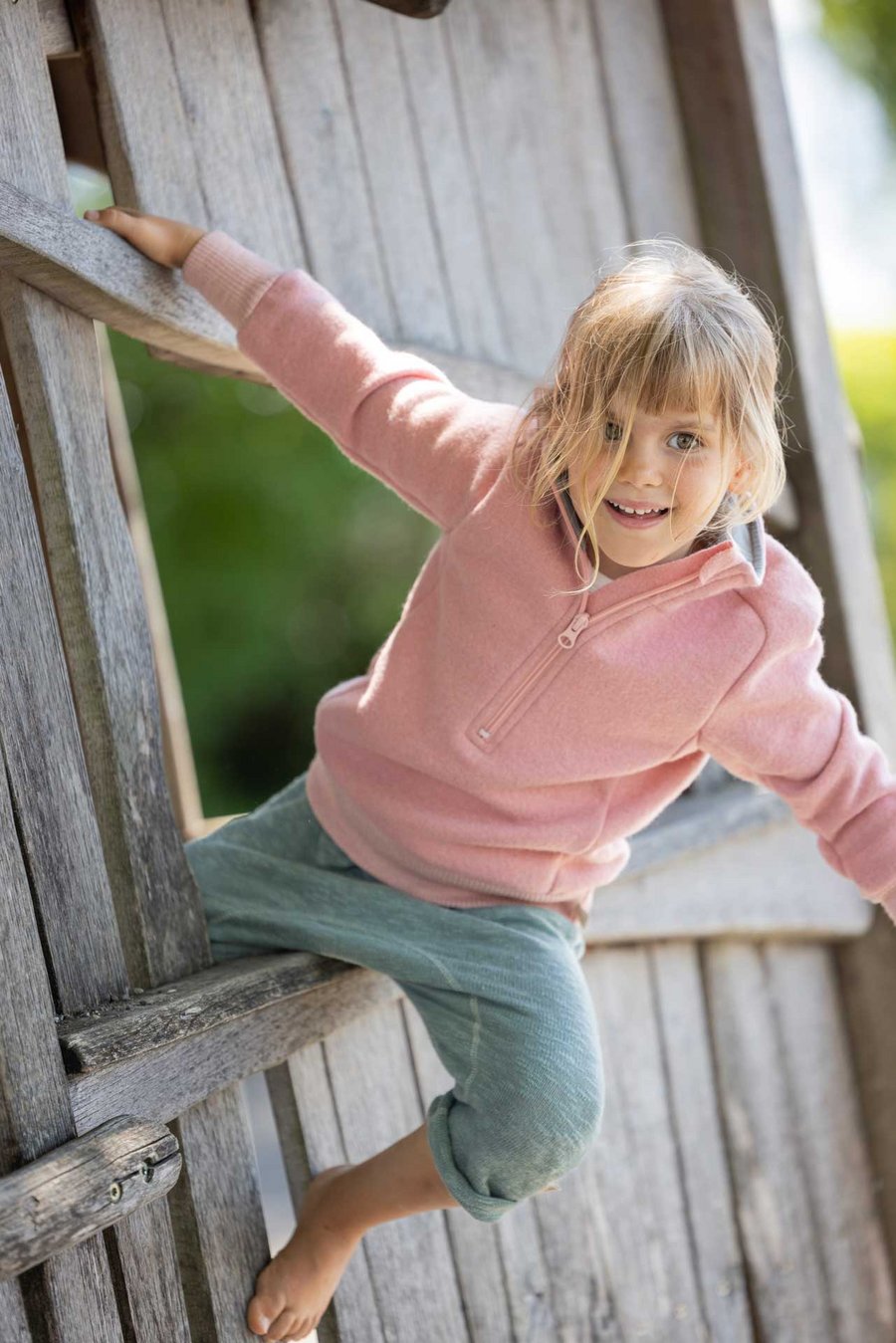 Mädchen im disana Troyer Softpink und Leinen-Strickhose in Jade sitzt auf dem Fenster einer Holzhütte auf einem Spielplatz