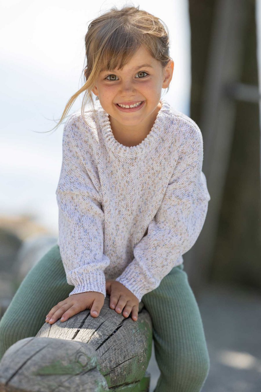 Mädchen mit goldbraunen Haaren in disana Baumwoll-Pullover "Spray", Natur-Spray, sitzt draußen lachend auf Holzspielzeug