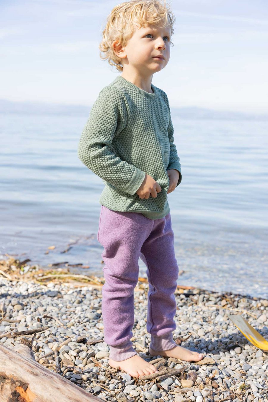 Blonder Junge im disana Wabenstrick-Pullover, Jade, und Pumphose, Flieder steht barfuß am Seeufer, 100% Bio-Merinowolle